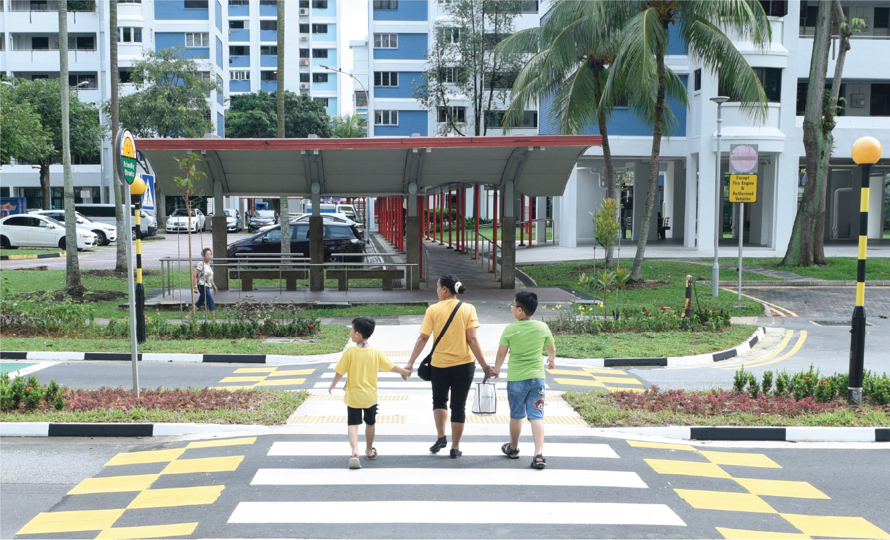 A woman holds the hands of two children while crossing a yellow and white crosswalk. Buildings and tropical trees are in the background.